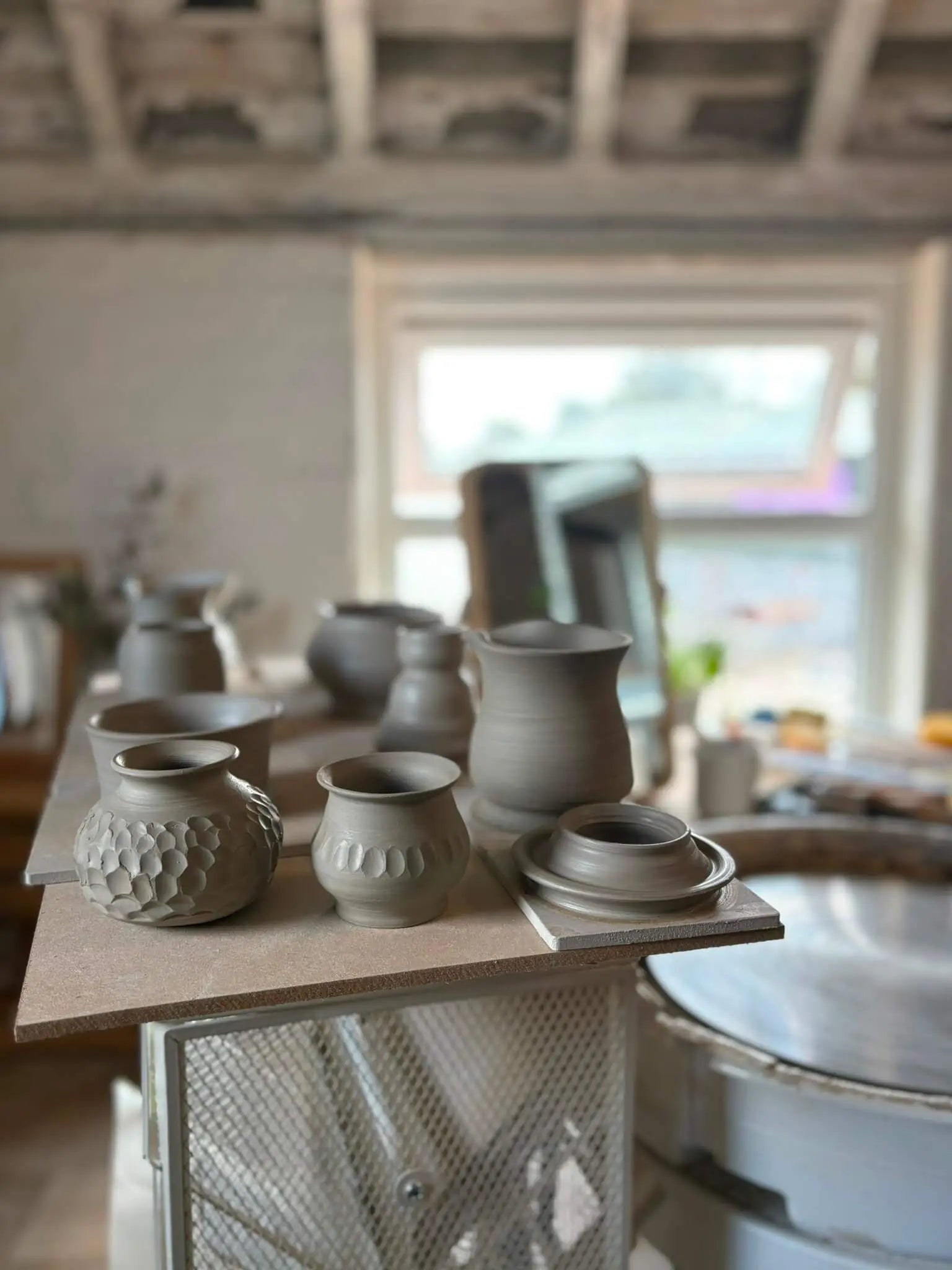 A group of handmade ceramics before the firing process sitting on top of a wooden table in a ceramics studio.