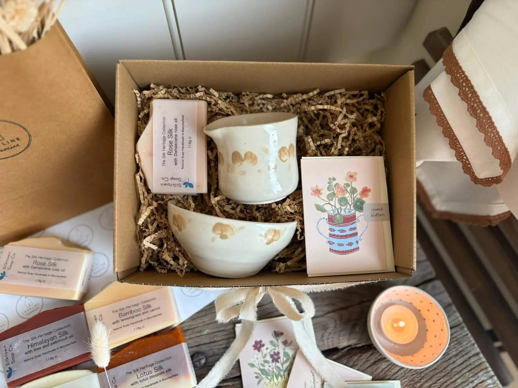 A curated gift box set containing a white ceramic tea set with gold accents, a small cream-colored soap, a floral card, and other tea accessories nestled in shredded paper inside a cardboard box.