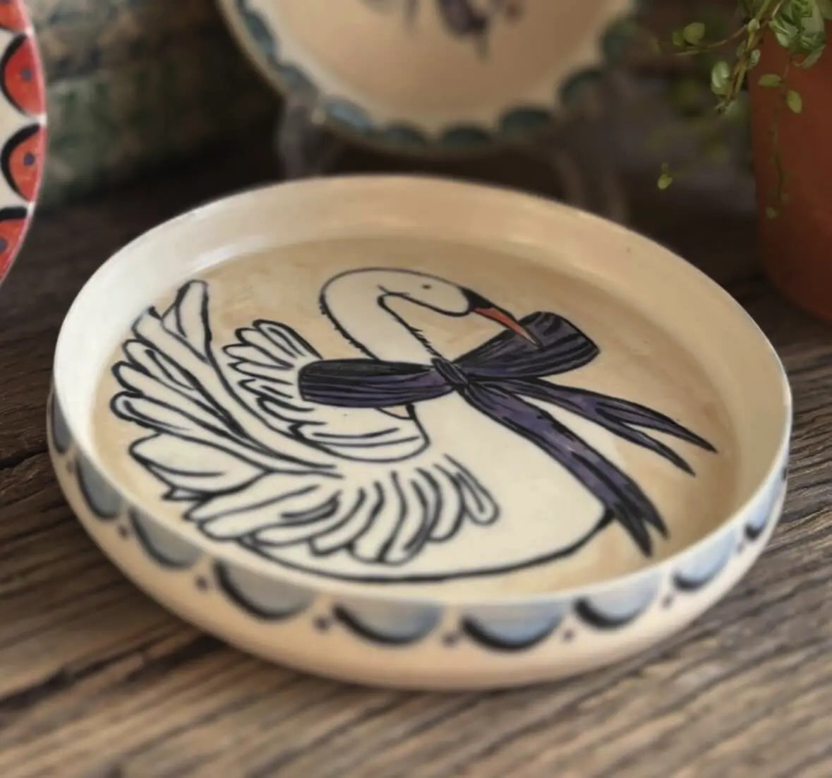 A cream ceramic trinket dish painted with a white swan wearing a purple bow, with a scalloped blue-and-black border, displayed on a wooden table.