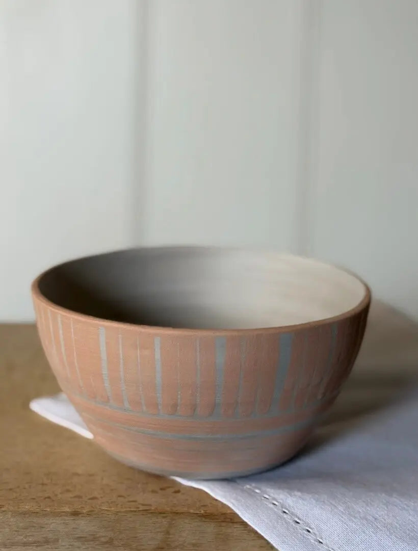A handmade ceramic bowl with a terracotta exterior featuring vertical ribbed stripes and a smooth, matte beige interior, resting on a white napkin on a wooden surface.
