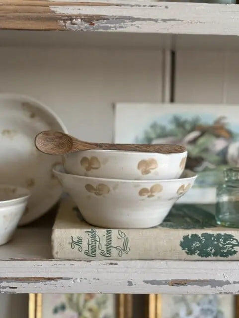 A stack of white ceramic bowls with gold floral motifs sits on a distressed white wooden shelf next to a wooden spoon, with vintage books and framed art in the background.