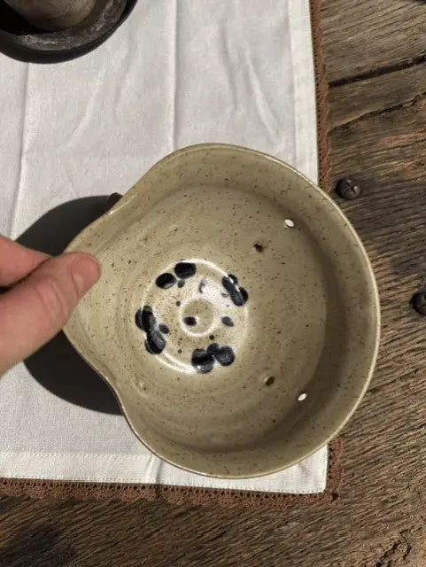 A handmade ceramic bowl with a speckled beige glaze and black spots inside, held by a hand over a white cloth on a rustic wooden table.