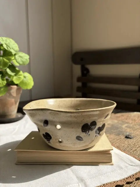 A handmade speckled beige ceramic bowl with black floral motifs and cutout holes, resting on a stack of books on a white tablecloth with a potted plant in the background.