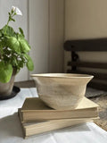 A handmade ceramic bowl with a speckled beige and cream glaze sits on top of two vintage hardcover books on a white tablecloth. A green potted plant is visible in the soft-focus background on the left.