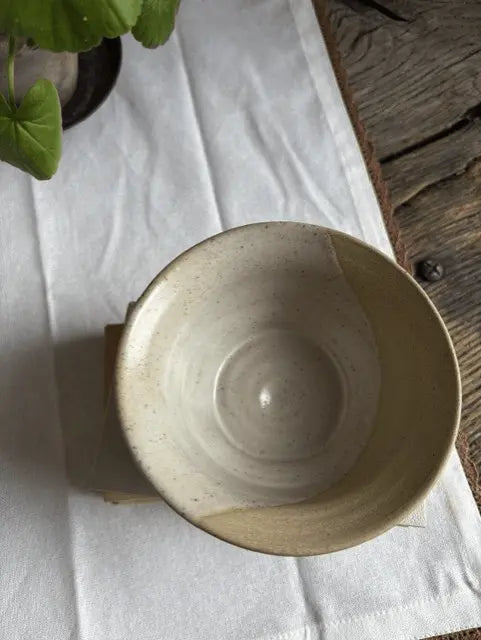 A handmade ceramic bowl with a speckled beige glaze, photographed from a top-down angle on a white fabric runner with a rustic wooden surface in the background.