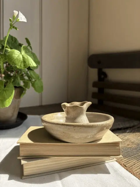 A small beige ceramic pitcher with a flared rim sits on top of two stacked books on a white tablecloth. A potted green plant is visible on the left, and a dark wooden chair is softly blurred in the background.