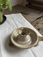 A small handmade ceramic vessel with a wide shallow base and a small central spout, shown on a white cloth over a rustic wooden surface.