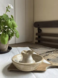 A small handmade ceramic pitcher and matching bowl set on a bamboo tray, placed on a white tablecloth in a softly lit room with a potted plant and wooden furniture in the background.