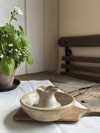 A small handmade ceramic pitcher and matching bowl set on a bamboo tray, placed on a white tablecloth in a softly lit room with a potted plant and wooden furniture in the background.