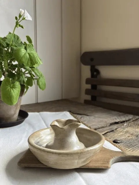 A small handmade ceramic pitcher and matching bowl set on a bamboo tray, placed on a white tablecloth in a softly lit room with a potted plant and wooden furniture in the background.