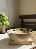 A handmade ceramic bowl with a speckled beige finish and black floral accents, resting on a stack of books on a white tablecloth in a sunlit room.
