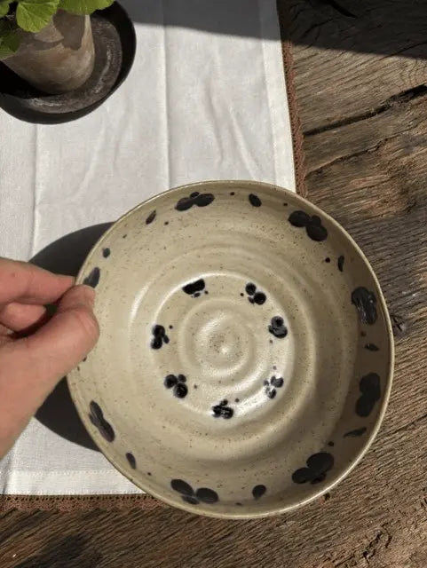 A handmade ceramic bowl with a speckled beige glaze and black hand-painted spots, held by a hand on a rustic wooden table with a white cloth.