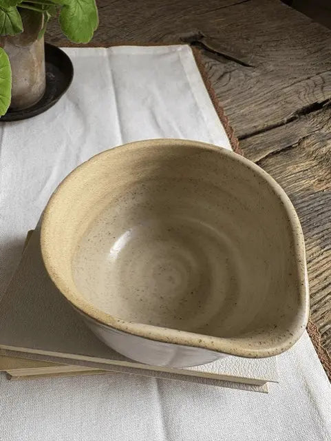 A handmade ceramic bowl with a wide, rounded shape and a speckled beige glaze, resting on a stack of books on a white linen cloth over a rustic wooden table.