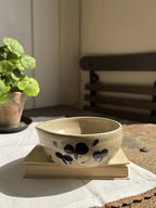 A handmade ceramic bowl with a beige speckled glaze and dark blue hand-painted floral motifs, resting on a vintage book on a white tablecloth in a sunlit room.