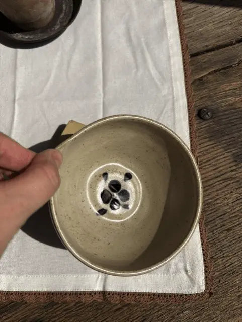 A hand holding a handmade ceramic bowl with a speckled beige interior and a black floral pattern on the bottom, set on a white cloth over a wooden surface.