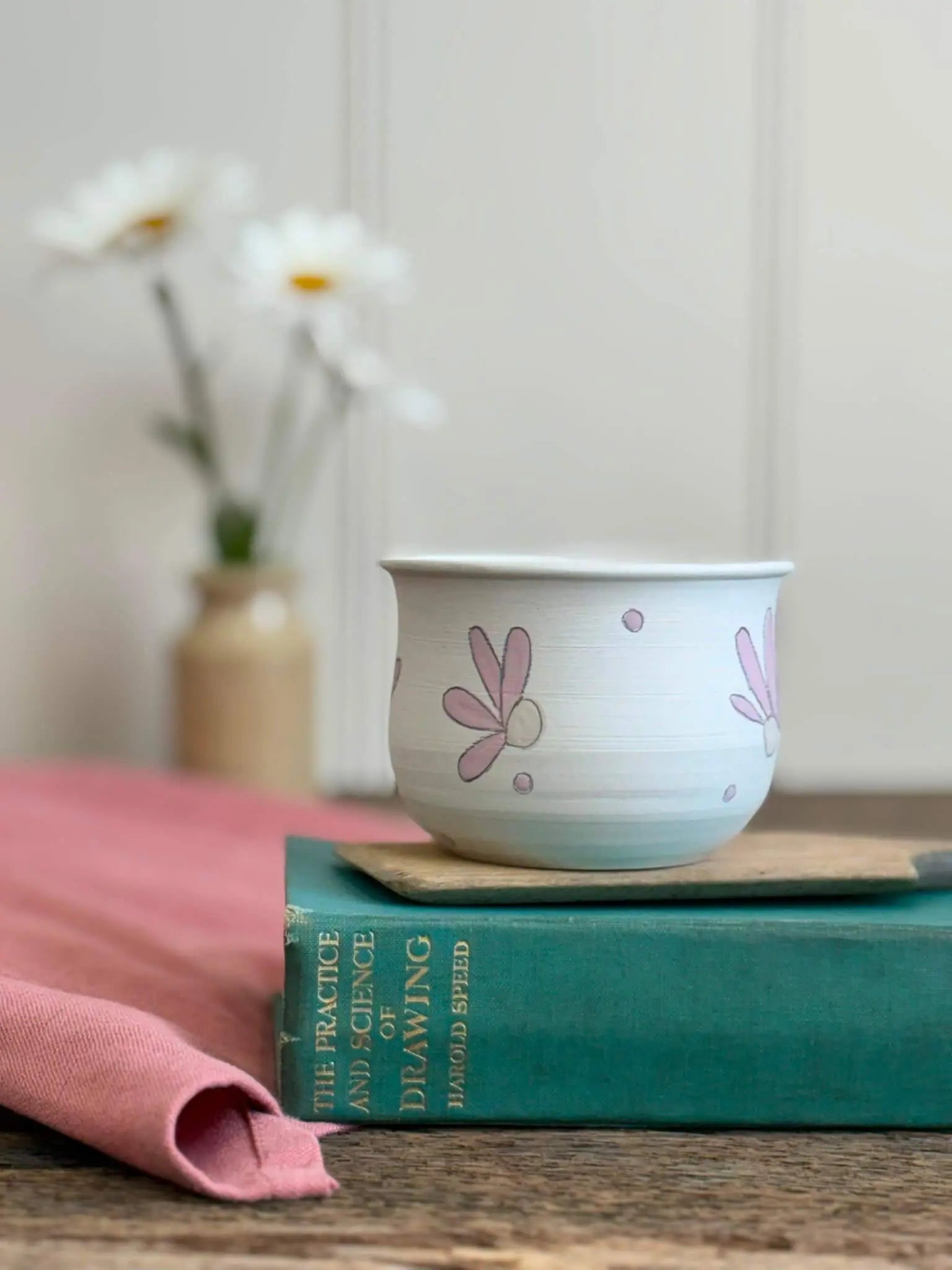 A white ceramic cup with pink floral illustrations sits on top of a teal book, with a pink cloth and blurred white flowers in the background.