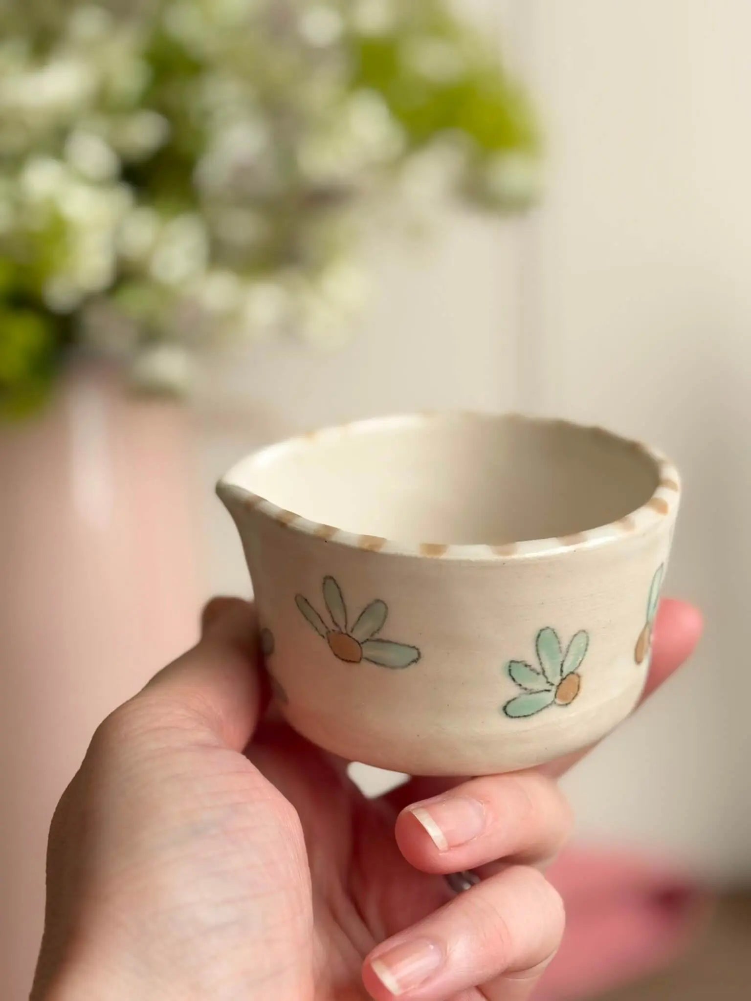 A hand holding a small handmade ceramic cup with a daisy pattern, set against a soft, blurred background of white flowers and green leaves.