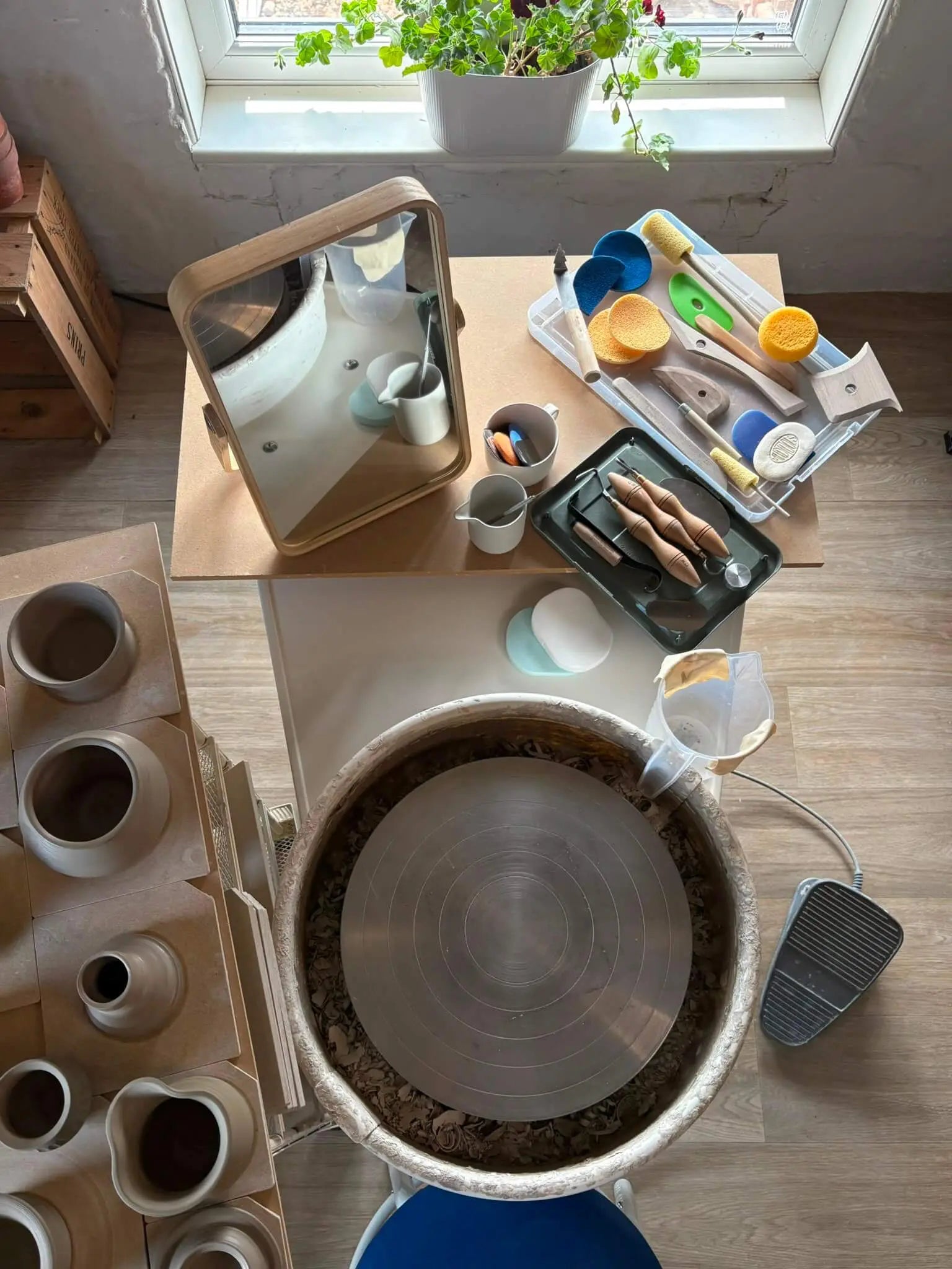 An overhead view of a pottery wheel workspace with a large spinning wheel in the center, multiple clay vessels and a small mirror on a table, tools in a tray, a foot pedal, and a potted plant by the window.