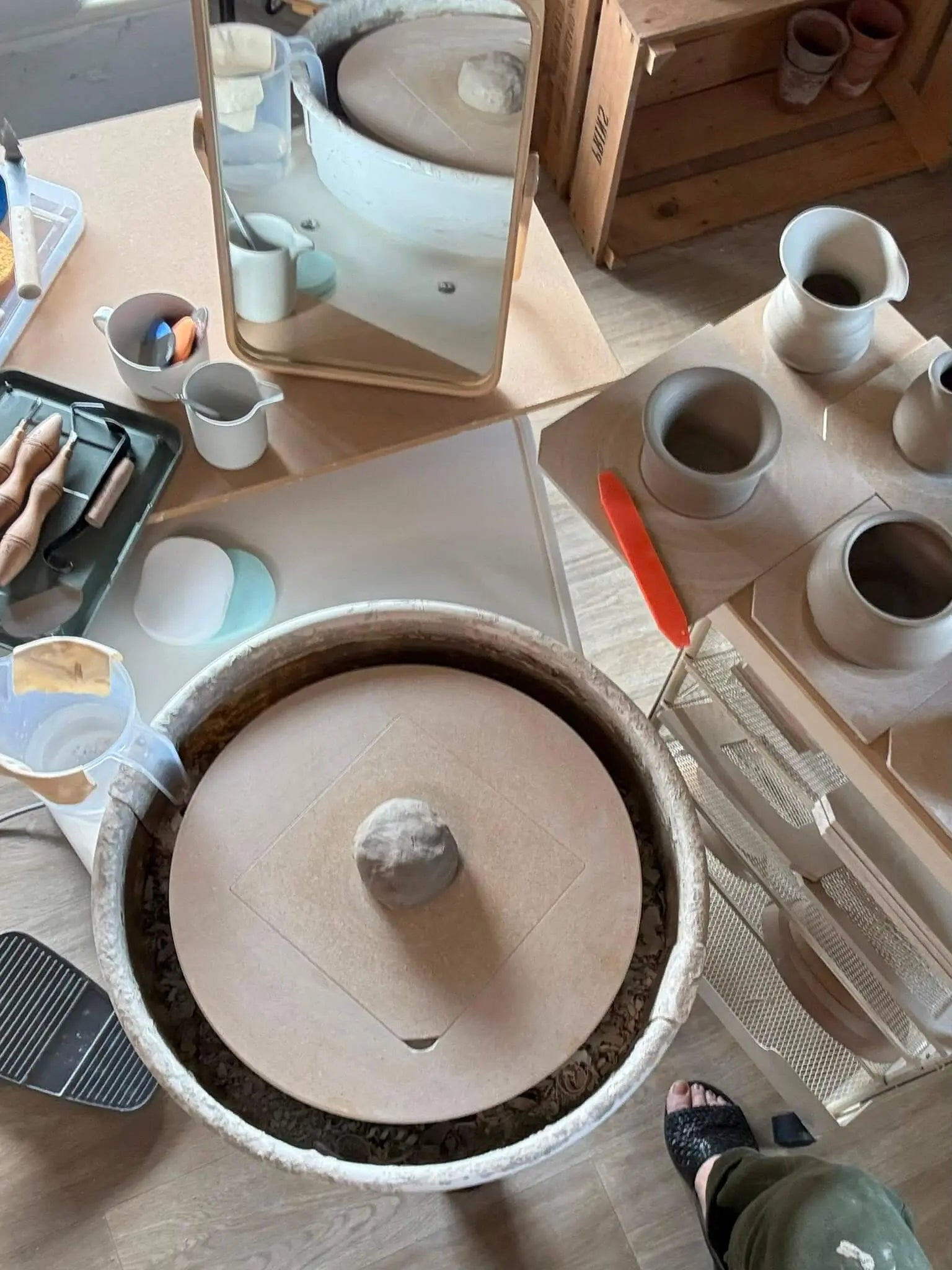 A pottery wheel and clay workbench in a studio, with a large round clay piece in the foreground, small ceramic vessels, tools, a mirror, and a person's foot visible at the bottom right.