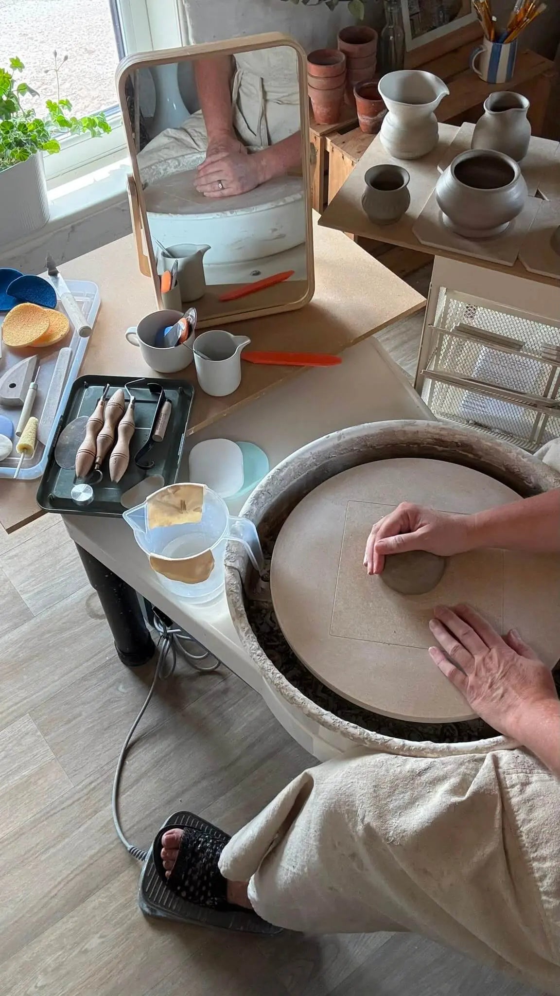 A person's hands and lower body are shown shaping a clay pot on a pottery wheel in a bright, wood-furnished pottery studio. Various clay tools, a mirror, and finished ceramic vessels are visible on the workbench.