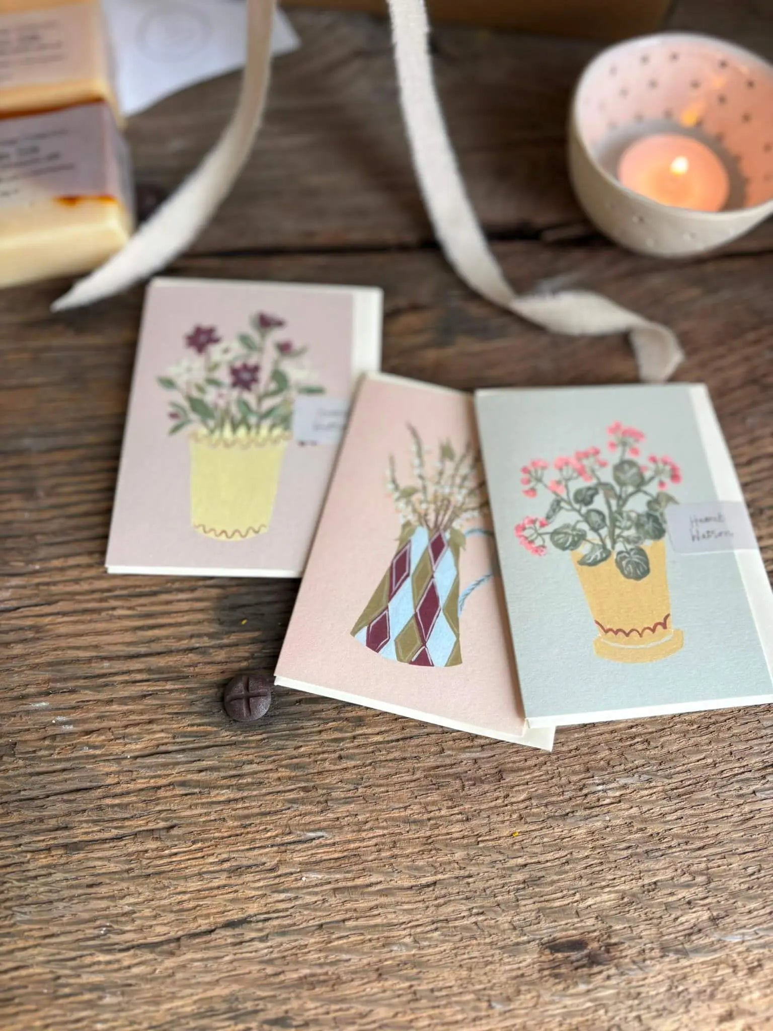 Three small illustrated cards or postcards with potted plant designs are arranged on a rustic wooden surface, with a candle holder and ribbon in the background.