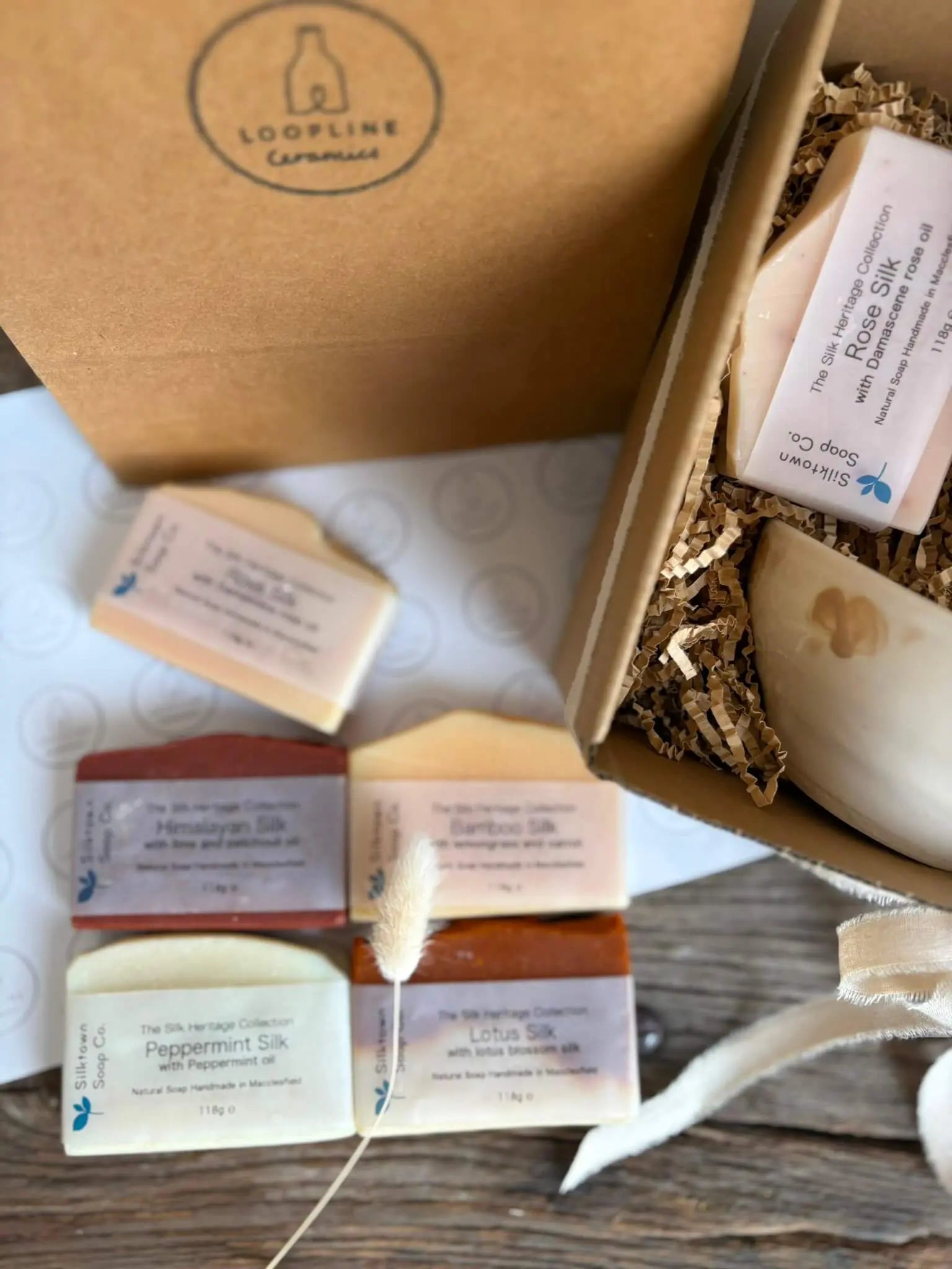 An open kraft gift box filled with packaged artisanal soaps, shown alongside several loose soap bars on a tabletop. The box lid has a visible Loopline Candles logo, and the soaps are wrapped in labels including Rice Silk, Bamboo Silk, and Lotus Silk.