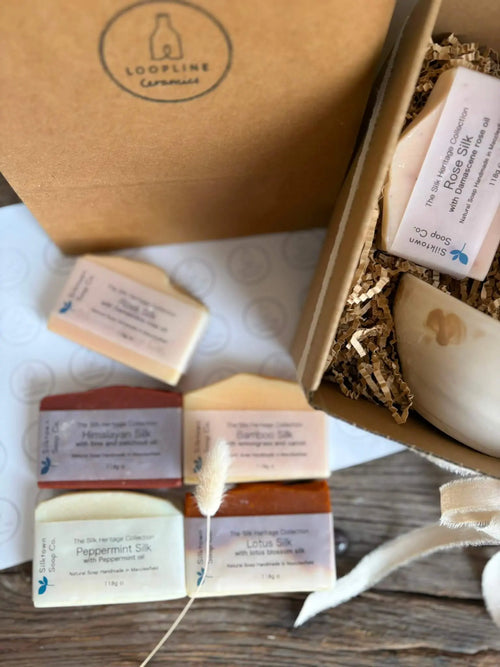 An open kraft gift box filled with packaged artisanal soaps, shown alongside several loose soap bars on a tabletop. The box lid has a visible Loopline Candles logo, and the soaps are wrapped in labels including Rice Silk, Bamboo Silk, and Lotus Silk.