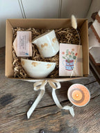 A gift box containing a cream-colored ceramic bowl, a matching cup, a small pink soap bar, a floral card, and a dried bunny tail, arranged in brown paper shred on a rustic wooden table.