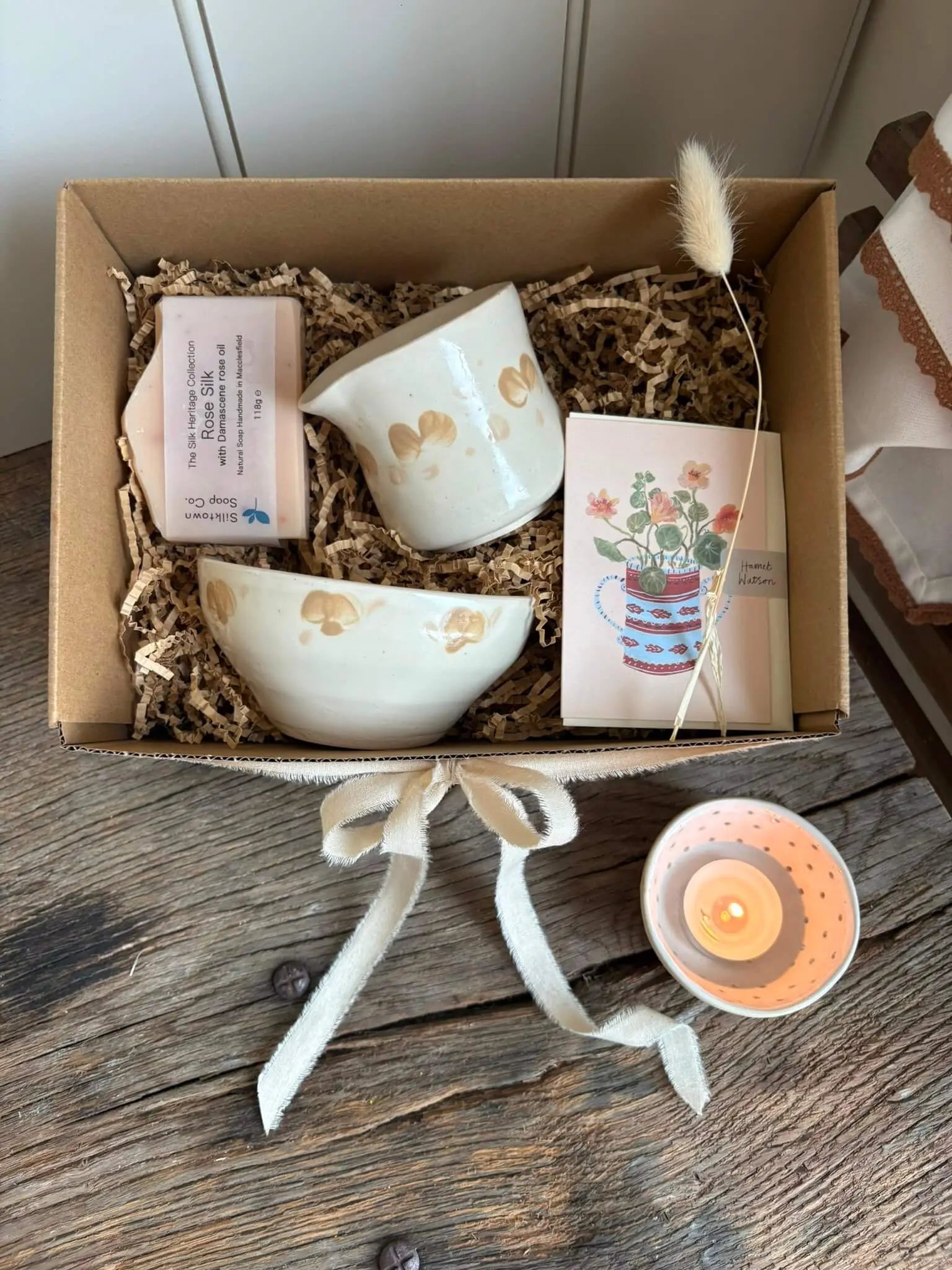 A gift box containing a cream-colored ceramic bowl, a matching cup, a small pink soap bar, a floral card, and a dried bunny tail, arranged in brown paper shred on a rustic wooden table.