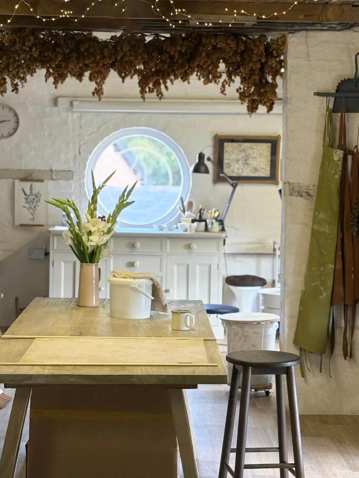 A rustic interior scene with a light wood table in the foreground, white cabinets, a round window, hanging dried botanicals, string lights, and a stool, suggesting a home or workshop environment.