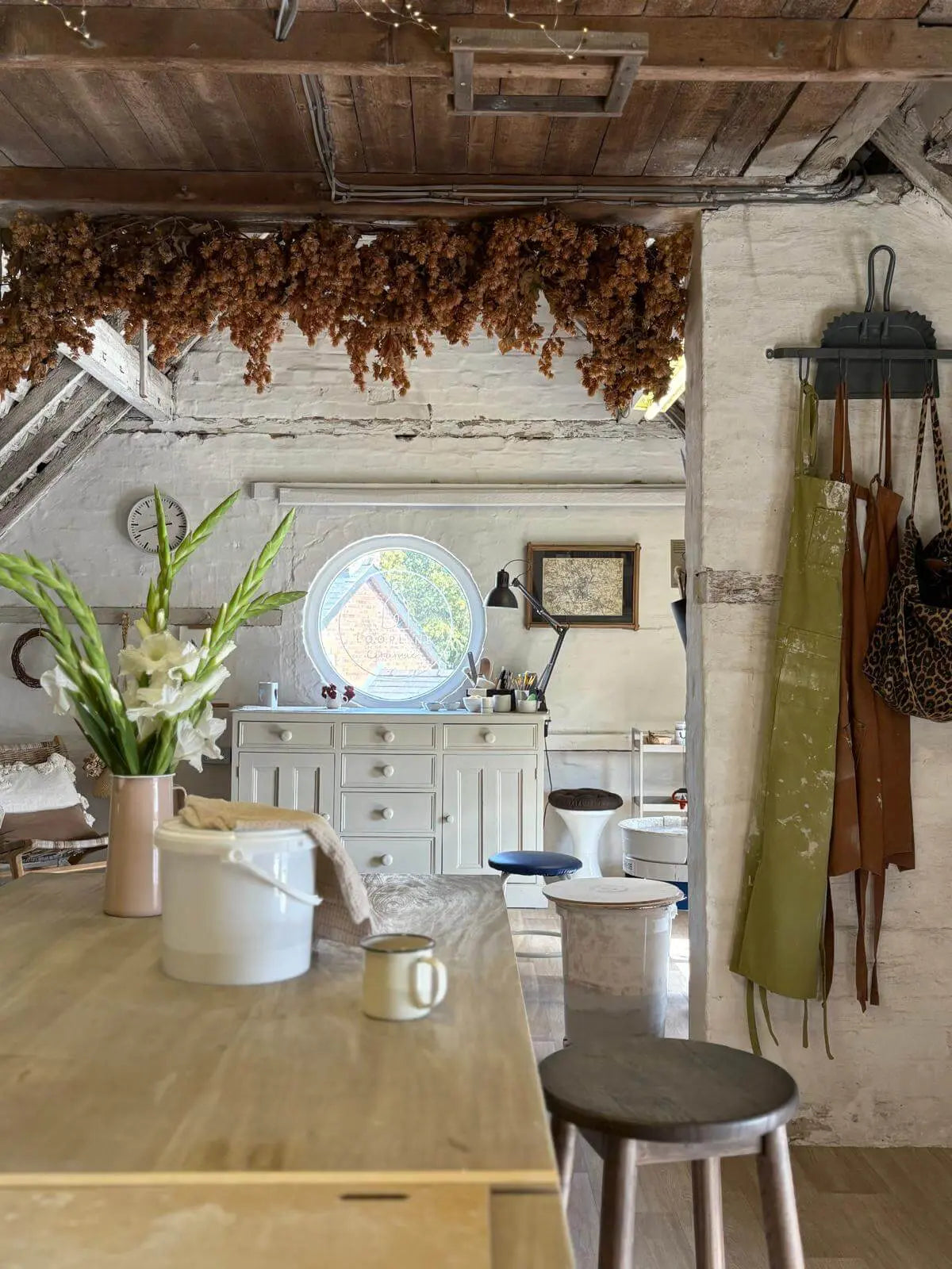 A rustic kitchen interior with a light wood table in the foreground, white brick walls, hanging aprons, dried brown botanicals draped across the ceiling, and a round window in the background.