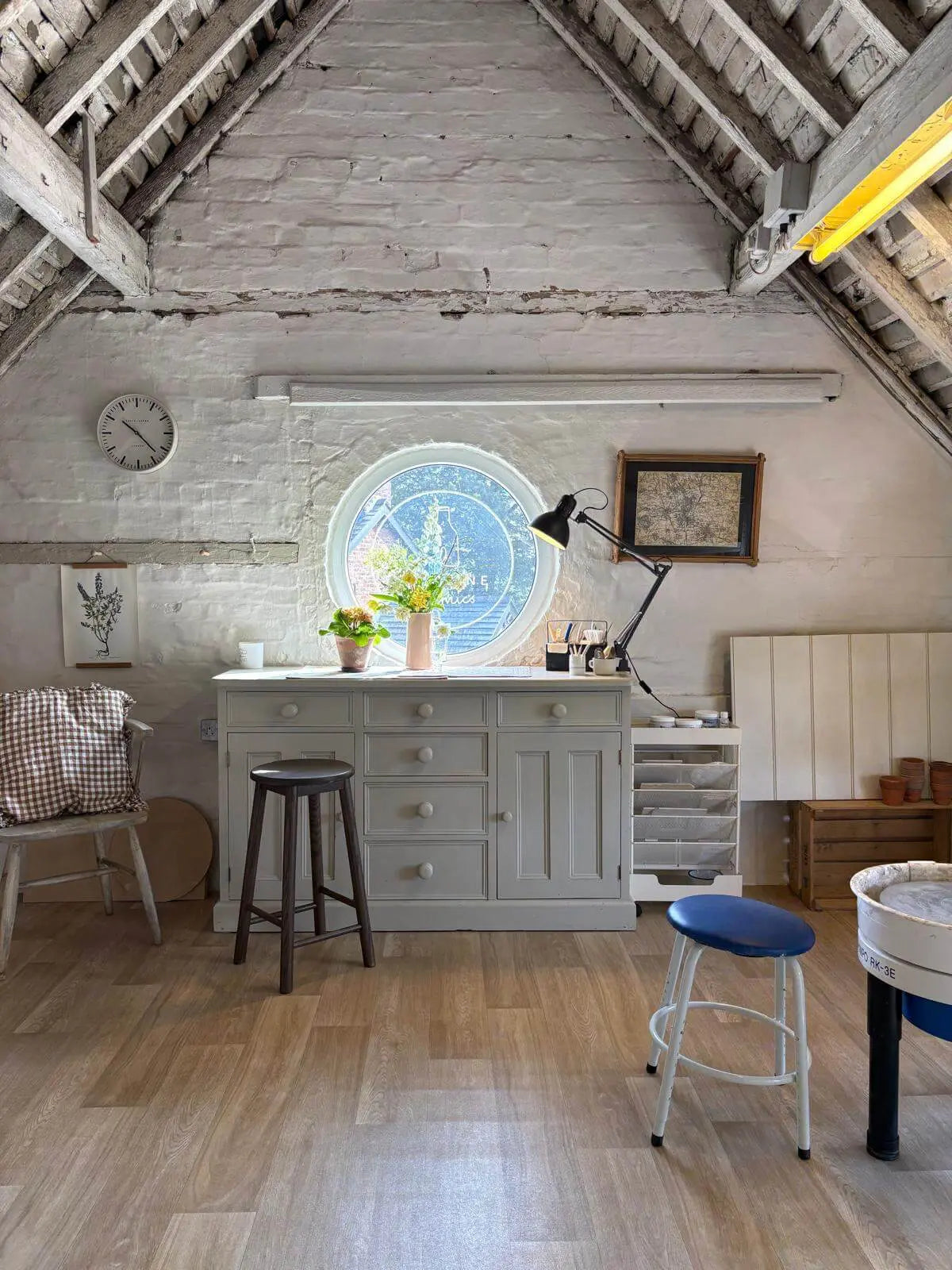 A rustic attic room with exposed wooden rafters and light wood floors, featuring a pale gray storage cabinet centered beneath a round window, with stools and wall decor in the room.