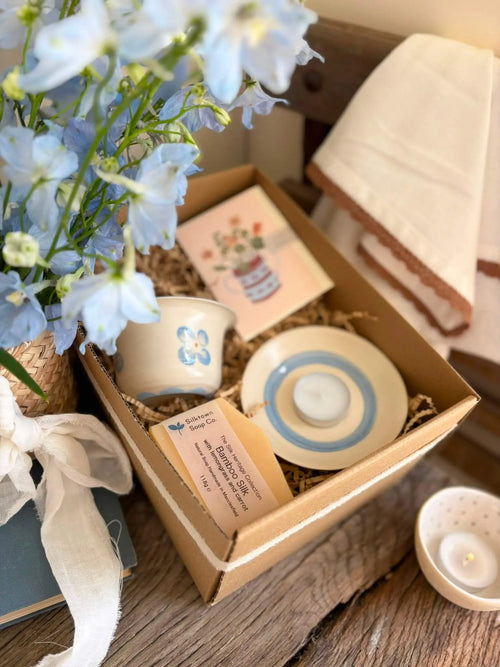 A gift box filled with small ceramic tea or candle holders, a floral card, and a card with text, styled on a wooden surface with blue flowers, a white ribbon, and a folded cream cloth nearby.