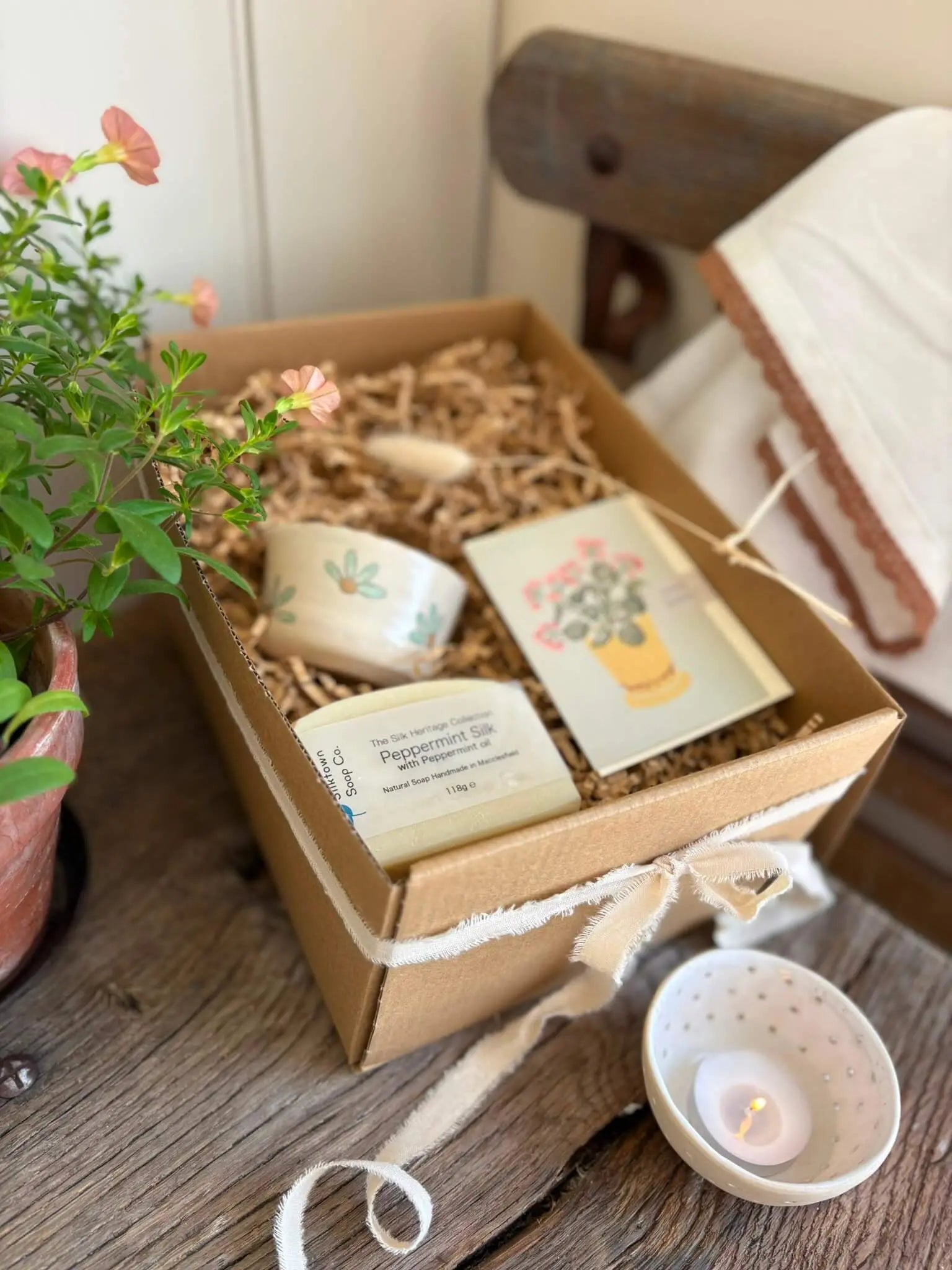 A gift box containing a small ceramic candle, a card with a floral illustration, and a bar of soap, arranged in a kraft box with shredded paper on a rustic wooden table next to a potted plant.