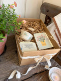 A handmade gift box containing a small ceramic cup, a candle, a card, and a soap, nestled in shredded paper inside a brown cardboard box tied with a white ribbon, set on a rustic wooden table next to a potted plant.