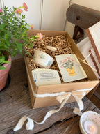 A handmade gift box containing a small ceramic cup, a candle, a card, and a soap, nestled in shredded paper inside a brown cardboard box tied with a white ribbon, set on a rustic wooden table next to a potted plant.