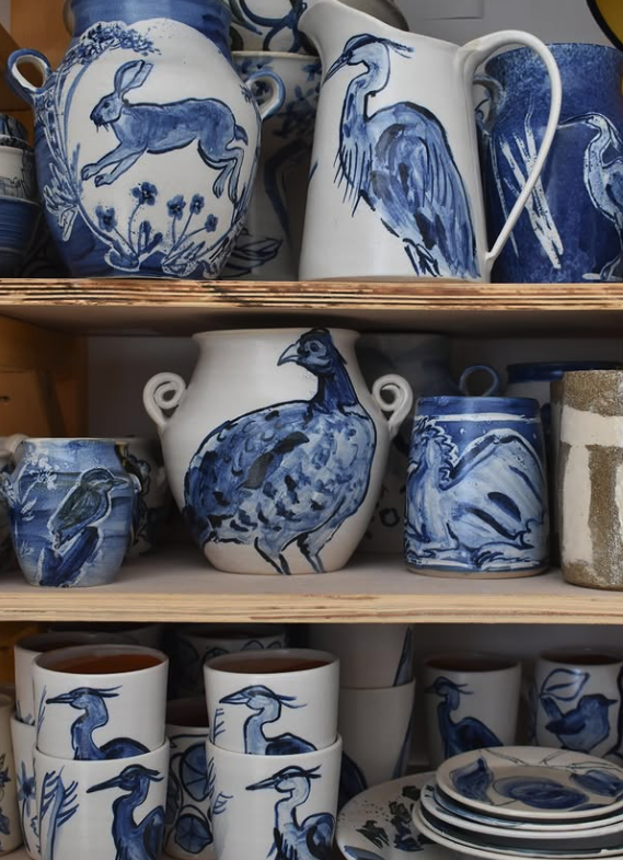 Blue and white ceramic tableware displayed on wooden shelves, including pitchers, cups, mugs, and plates with bird and rabbit motifs.