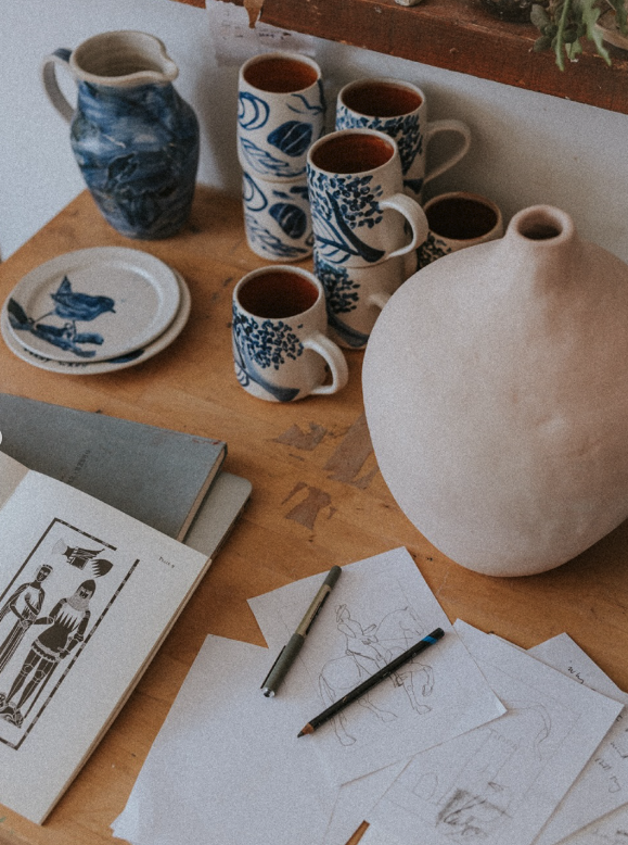 A styled tabletop arrangement of ceramic pottery and mugs on a wooden surface, including a large matte beige vase, blue-and-white patterned cups and plates, a blue-and-white pitcher, sketch papers, pencils, and an open notebook.