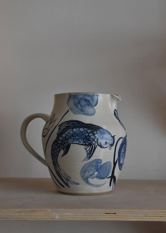 A handmade ceramic pitcher with a blue and white fish pattern, resting on a light wooden shelf against a neutral gray background.