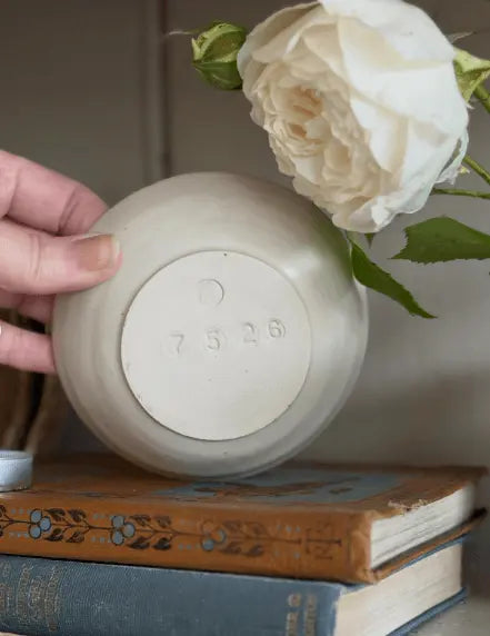 A hand holds the bottom of a matte off-white ceramic vase or planter, showing the stamped number '7526' on the base. The vase is styled on top of two vintage books with a large white rose resting behind it.