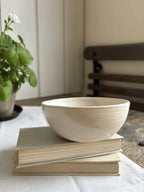 A small off-white ceramic bowl with a textured, ribbed exterior and smooth interior, resting on a stack of two books on a white tablecloth. A potted green plant is visible in the soft-focus background on the left.
