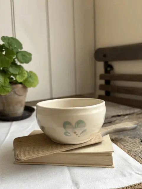 A small cream-colored ceramic bowl with a light green bunny face design sits on a stack of vintage books on a white cloth, with a potted plant and a wooden chair blurred in the background.