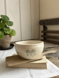 A small cream-colored ceramic bowl with a light green bunny face design sits on a stack of vintage books on a white cloth, with a potted plant and a wooden chair blurred in the background.