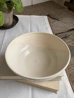 A large, shallow ceramic bowl in a warm cream or off-white glaze, resting on a stack of books on a white tablecloth over a rustic wooden table.