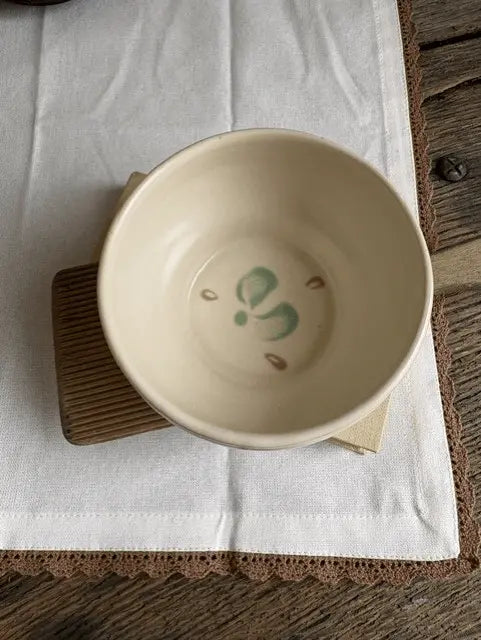 A handmade ceramic bowl with a hand-painted green and brown floral design inside, resting on a wooden coaster over a white linen cloth on a rustic wooden table.