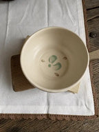 A handmade ceramic bowl with a hand-painted green and brown floral design inside, resting on a wooden coaster over a white linen cloth on a rustic wooden table.