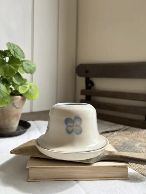 A small white ceramic cup with a blue floral design, shown upside down on top of a wooden spoon and a book in a softly lit indoor setting.