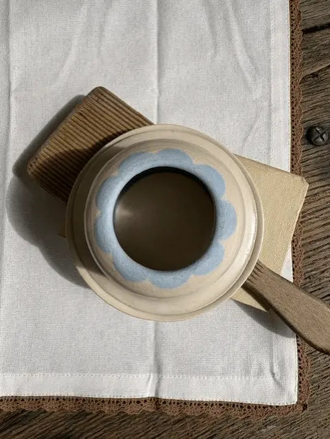 A ceramic cup with a cream-colored exterior, a light blue scalloped rim, and a dark brown interior, resting on a white cloth over a wooden surface.