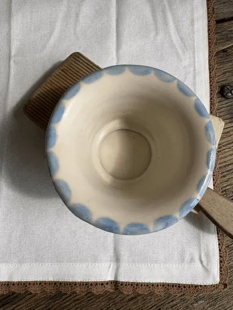 A handmade ceramic bowl with a scalloped blue rim, resting on a white linen cloth and a brown wooden block over a rustic wooden table.