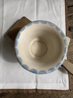 A handmade ceramic bowl with a scalloped blue rim, resting on a white linen cloth and a brown wooden block over a rustic wooden table.